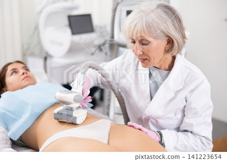 Elderly female doctor doing Rf lifting procedure on belly of young woman in beauty salon. Treatment of flabby skin Elderly female doctor doing Rf lifting procedure on belly of young woman in beauty salon. Treatment of flabby skin 141223654