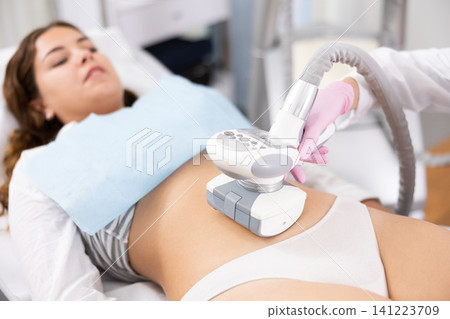 Young woman getting ultrasound cavitation body contouring treatment, anti-cellulite therapy, vacuum massage from unrecognizable cosmetologist. Hardware cosmetology for body sculpting Young woman getting ultrasound cavitation body contouring treatment, anti-cellulite therapy, vacuum massage from unrecognizable cosmetologist. Hardware cosmetology for body sculpting 141223709
