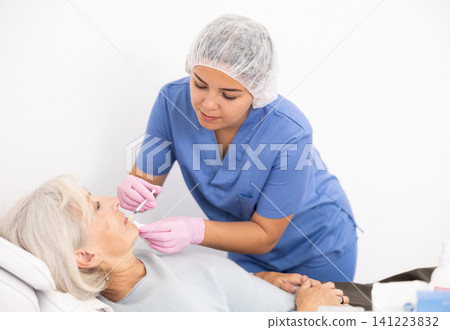 Young woman cosmetologist in uniform making injections for lip augmentation and anti-wrinkle of elderly female in esthetic clinic indoors Young woman cosmetologist in uniform making injections for lip augmentation and anti-wrinkle of elderly female in esthetic clinic indoors 141223832
