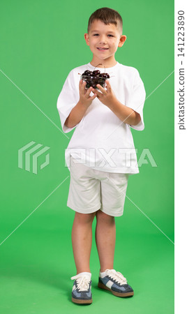 Boy in white t-shirt and shorts happily eats ripe cherries on green background 141223890