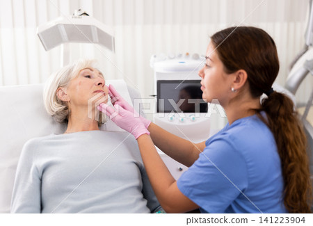 Young female cosmetologist in gloves examining face skin of elderly female client before beauty procedure in spa center indoors Young female cosmetologist in gloves examining face skin of elderly female client before beauty procedure in spa center indoors 141223904
