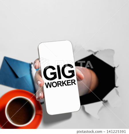 A hand holding a smartphone displaying the text 'GIG WORKER' through a torn paper background A hand holding a smartphone displaying the text 'GIG WORKER' through a torn paper background 141225391