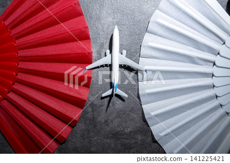 A top-down view of a small model airplane placed in the center between a vibrant red folding fan and a patterned white fan on a textured gray background, symbolizing travel and choices A top-down view of a small model airplane placed in the center between a vibrant red folding fan and a patterned white fan on a textured gray background, symbolizing travel and choices 141225421