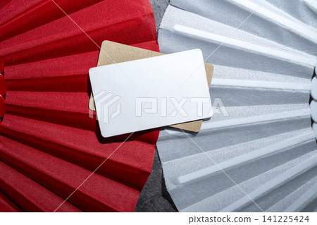 A close-up top view of a blank white rectangular card placed on top of a cork surface, framed by textured red and white paper fan pleats, perfect for design mockups A close-up top view of a blank white rectangular card placed on top of a cork surface, framed by textured red and white paper fan pleats, perfect for design mockups 141225424