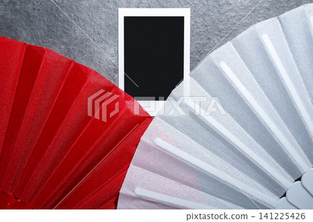 A close-up view of a red fabric fan and a white lace fan separated by a blank polaroid-style instant photo frame against a dark textured stone background A close-up view of a red fabric fan and a white lace fan separated by a blank polaroid-style instant photo frame against a dark textured stone background 141225426