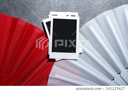 Top view of three blank Polaroid-style photo frames arranged between vibrant red and white accordion hand fans on a dark textured background. Perfect for creative project mockups Top view of three blank Polaroid-style photo frames arranged between vibrant red and white accordion hand fans on a dark textured background. Perfect for creative project mockups 141225427