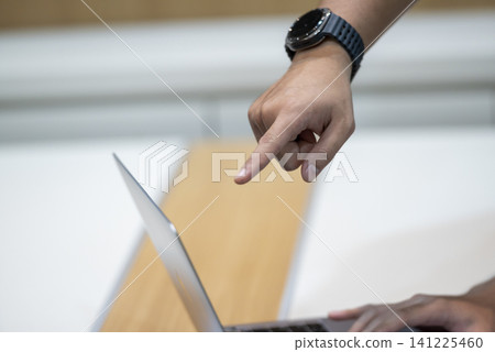A close-up shot of a person's hand pointing with an index finger at a laptop screen. The person is wearing a black smartwatch and working on a desk A close-up shot of a person's hand pointing with an index finger at a laptop screen. The person is wearing a black smartwatch and working on a desk 141225460