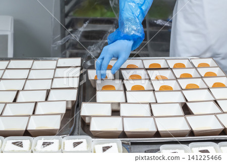 Food factory worker in blue sterile gloves arranging orange gelatin desserts into small square paper containers on a tray in a professional kitchen 141225466