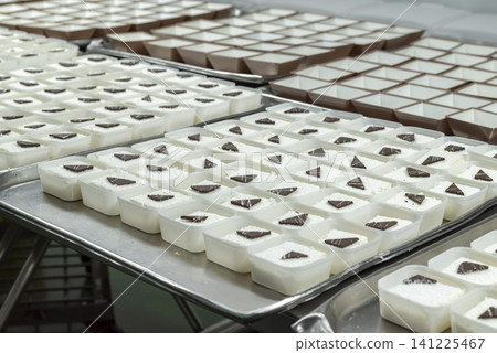 Trays of mass-produced white cream desserts topped with chocolate pieces, prepared in a commercial kitchen or catering facility Trays of mass-produced white cream desserts topped with chocolate pieces, prepared in a commercial kitchen or catering facility 141225467