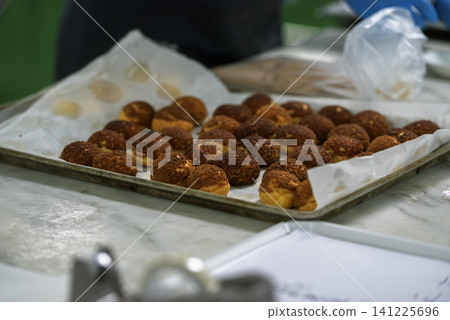 A metal tray lined with parchment paper holds rows of freshly baked golden-brown choux pastries with craquelin tops in a professional bakery kitchen 141225696