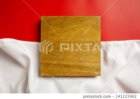 A top-down view of a square, textured wooden board positioned on white draped fabric against a vivid red background, ideal for product mockups and minimalist design layouts A top-down view of a square, textured wooden board positioned on white draped fabric against a vivid red background, ideal for product mockups and minimalist design layouts 141225902