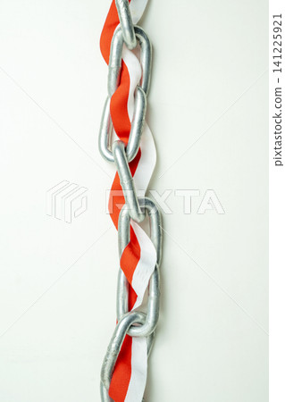 A close-up vertical shot of a silver metal chain intertwined with a red and white striped ribbon, symbolizing struggle, patriotism, or political bondage against a light background A close-up vertical shot of a silver metal chain intertwined with a red and white striped ribbon, symbolizing struggle, patriotism, or political bondage against a light background 141225921