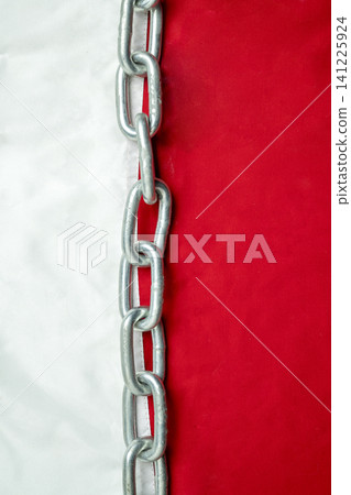 A close-up vertical shot of a strong steel chain dividing a vibrant red and textured white background, representing strength, division, or connection A close-up vertical shot of a strong steel chain dividing a vibrant red and textured white background, representing strength, division, or connection 141225924
