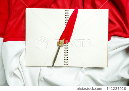 A high-angle studio shot featuring an elegant red quill pen resting on a blank spiral-bound notebook, set against the red and white fabric backdrop of the Indonesian national flag A high-angle studio shot featuring an elegant red quill pen resting on a blank spiral-bound notebook, set against the red and white fabric backdrop of the Indonesian national flag 141225928