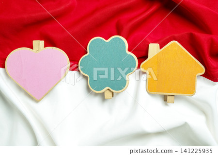 Three colorful wooden clips in heart, tree, and house shapes arranged on a draped Indonesian red and white national flag background Three colorful wooden clips in heart, tree, and house shapes arranged on a draped Indonesian red and white national flag background 141225935