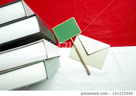 A stack of books, an envelope, and a blank green wooden sign on the red and white flag of Indonesia. Great for educational, national holiday, or patriotic concepts A stack of books, an envelope, and a blank green wooden sign on the red and white flag of Indonesia. Great for educational, national holiday, or patriotic concepts 141225940