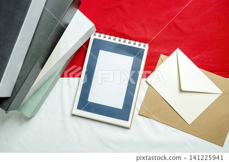 A flat lay composition featuring a spiral notebook, envelopes, and stacked books arranged on a red and white textured fabric background A flat lay composition featuring a spiral notebook, envelopes, and stacked books arranged on a red and white textured fabric background 141225941