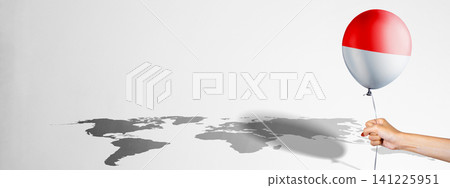 A conceptual image showing a hand holding a balloon with the Indonesian flag, casting a shadow of a world map on a plain background, representing international relations, travel, or global influence 141225951