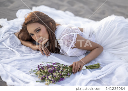 A stunning young Indonesian southeast asian bride in a white lace wedding dress lying on a white cloth on the sand, holding a beautiful bouquet of wildflowers and looking at the camera A stunning young Indonesian southeast asian bride in a white lace wedding dress lying on a white cloth on the sand, holding a beautiful bouquet of wildflowers and looking at the camera 141226084