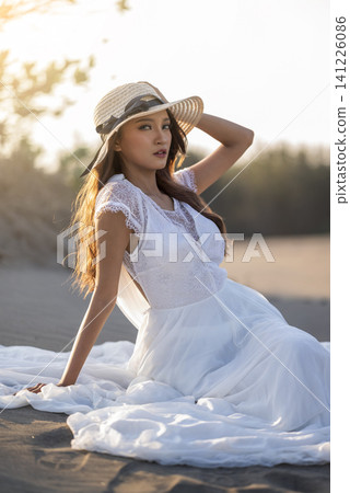 A beautiful young Indonesian southeast asian woman in an elegant white lace dress and straw hat poses gracefully on the sandy beach during golden hour A beautiful young Indonesian southeast asian woman in an elegant white lace dress and straw hat poses gracefully on the sandy beach during golden hour 141226086