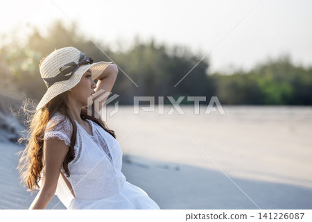 A beautiful young Indonesian southeast asian woman in an elegant white lace dress and straw hat poses gracefully on the sandy beach during golden hour 141226087