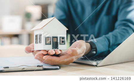 Real estate contract for property mortgage finance approved, home insurance document protects homeowner and house tenant, investment strategy involves agent holding model house near laptop Real estate contract for property mortgage finance approved, home insurance document protects homeowner and house tenant, investment strategy involves agent holding model house near laptop 141226169