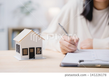 Close up of woman hand writing estate property residential investment agreement, signing domestic construction insurance mortgage contract with small house model on table 141226178