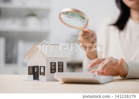 Woman holding magnifying glass, looking at house model on desk while using calculator to analyze finance, search for perfect home loan or plan real estate investment budget Woman holding magnifying glass, looking at house model on desk while using calculator to analyze finance, search for perfect home loan or plan real estate investment budget 141226179