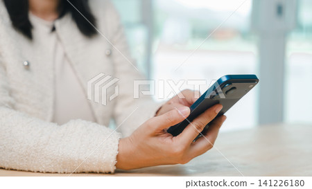 Woman uses mobile phone, smartphone technology online to connect, as she sits at wooden table wearing white textured jacket to browse social media or text friends in bright office space Woman uses mobile phone, smartphone technology online to connect, as she sits at wooden table wearing white textured jacket to browse social media or text friends in bright office space 141226180