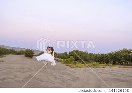 A Indonesian southeast asian romantic couple dressed in white strolling across sandy dunes during a soft pastel sunset, creating a serene and timeless atmosphere perfect for wedding or engagement A Indonesian southeast asian romantic couple dressed in white strolling across sandy dunes during a soft pastel sunset, creating a serene and timeless atmosphere perfect for wedding or engagement 141226198