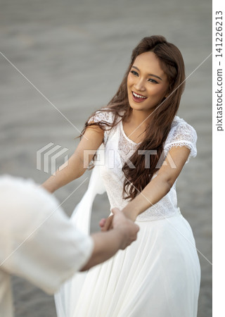 A happy young Indonesian southeast asian woman in a white dress holds hands and dances with her partner on a sandy beach during a romantic pre-wedding or engagement shoot A happy young Indonesian southeast asian woman in a white dress holds hands and dances with her partner on a sandy beach during a romantic pre-wedding or engagement shoot 141226213