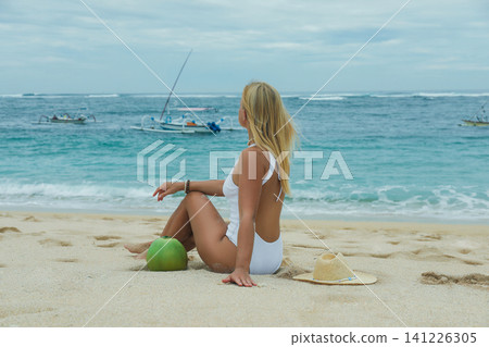 Woman on the beach enjoys a coconut drink, gazing at sea Woman on the beach enjoys a coconut drink, gazing at sea 141226305