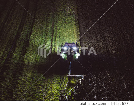 Aerial view from a drone Tractor is cultivating field at night. Tractor at night with long-distance lights from above.Night shifts Aerial view from a drone Tractor is cultivating field at night. Tractor at night with long-distance lights from above.Night shifts 141226773