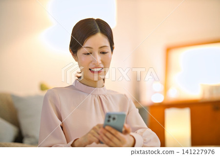 一位四十多歲的女性晚上在房間裡放鬆,看著她的智慧型手機。 一位四十多歲的女性晚上在房間裡放鬆,看著她的智慧型手機。 141227976