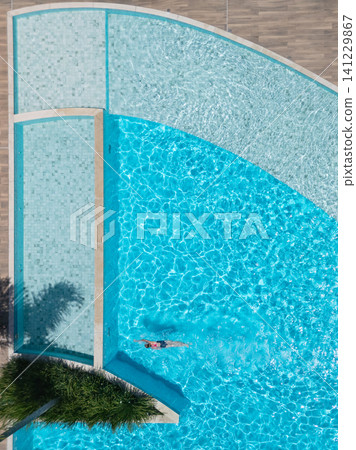 Young girl swimming in clear blue pool during sunny summer vacation. This aerial view captures family, friends, fun, and recreation at resort, relaxation and joyful outdoor moments. Young girl swimming in clear blue pool during sunny summer vacation. This aerial view captures family, friends, fun, and recreation at resort, relaxation and joyful outdoor moments. 141229867