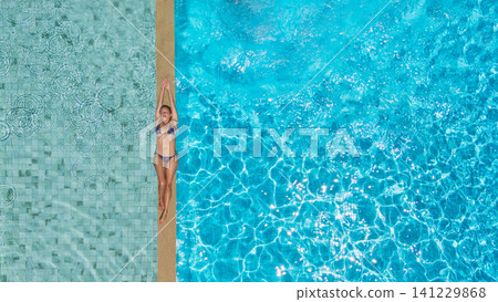 Young woman relax at the edge of clear blue pool during sunny summer vacation. This aerial view captures family,  friends, fun, and recreation at resort, relaxation and joyful outdoor moments. 141229868