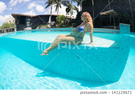 Pregnant woman relaxes in the shallow area of a swimming pool, enjoying warm sunshine and clear water. Summer vacation, healthy pregnancy, wellness and peaceful leisure in a tropical resort. Pregnant woman relaxes in the shallow area of a swimming pool, enjoying warm sunshine and clear water. Summer vacation, healthy pregnancy, wellness and peaceful leisure in a tropical resort. 141229871