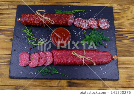 Salami sausages with ketchup and spices on black slate board on a wooden table Salami sausages with ketchup and spices on black slate board on a wooden table 141230276