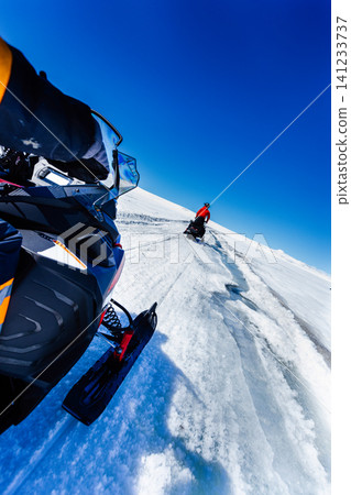 Close-up perspective of a snowmobile rider crossing a pristine Icelandic glacier, following another snowmobile across untouched snow beneath a vivid blue sky during a winter adventure. Close-up perspective of a snowmobile rider crossing a pristine Icelandic glacier, following another snowmobile across untouched snow beneath a vivid blue sky during a winter adventure. 141233737