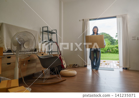 Smiling Asian woman carrying cardboard box through doorway on moving day Smiling Asian woman carrying cardboard box through doorway on moving day 141235071