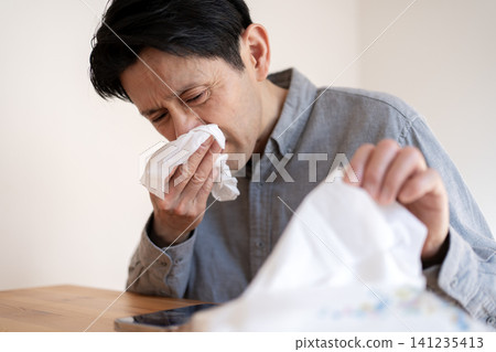 A man in his 40s, suffering from severe hay fever symptoms, blows his nose with a tissue. A man in his 40s, suffering from severe hay fever symptoms, blows his nose with a tissue. 141235413