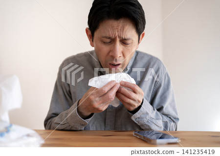 A man in his 40s, suffering from severe hay fever symptoms, blows his nose with a tissue. A man in his 40s, suffering from severe hay fever symptoms, blows his nose with a tissue. 141235419