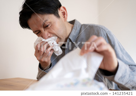 A man in his 40s, suffering from severe hay fever symptoms, blows his nose with a tissue. A man in his 40s, suffering from severe hay fever symptoms, blows his nose with a tissue. 141235421