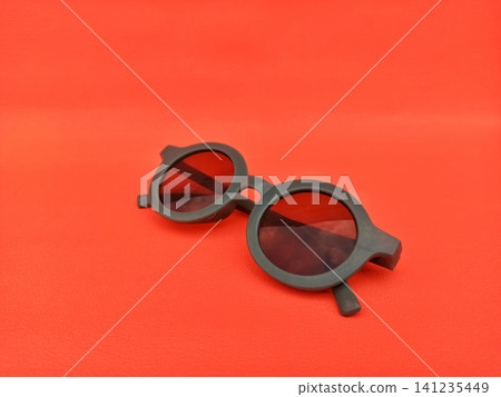 Stylish Round Sunglasses with Dark Lenses on Vibrant Red Background Stylish Round Sunglasses with Dark Lenses on Vibrant Red Background 141235449