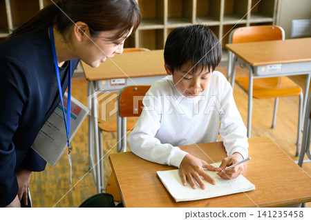 A female teacher gently watches over and provides individual instruction to a male elementary school student studying at his desk in the classroom. A female teacher gently watches over and provides individual instruction to a male elementary school student studying at his desk in the classroom. 141235458