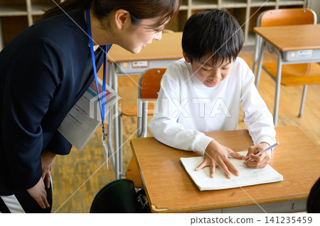 A female teacher gently watches over and provides individual instruction to a male elementary school student studying at his desk in the classroom. A female teacher gently watches over and provides individual instruction to a male elementary school student studying at his desk in the classroom. 141235459