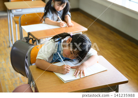 A female elementary school student smiles as she looks at her notebook while receiving individual tutoring from a female teacher in the classroom. 141235988