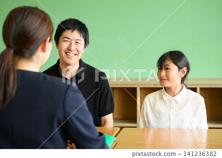 A female teacher, a father, and an elementary school girl are having a friendly and cheerful three-way meeting in a classroom (career guidance/tutoring). 141236382