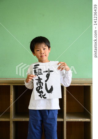 A male elementary school student proudly shows off his calligraphy artwork in the classroom, which has the word "sushi" written on it. A male elementary school student proudly shows off his calligraphy artwork in the classroom, which has the word "sushi" written on it. 141236439