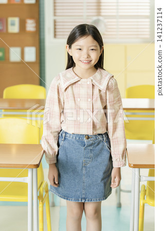 Smiling Asian schoolgirl posing with a backpack between classroom desks Smiling Asian schoolgirl posing with a backpack between classroom desks 141237114